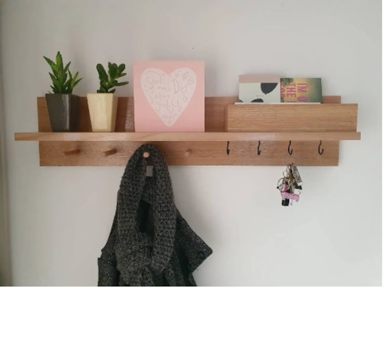 May include: A wooden wall shelf with a light brown finish, featuring a top shelf with small potted plants and a pink square with white text. Below are hooks for hanging items, with a scarf and keys visible.