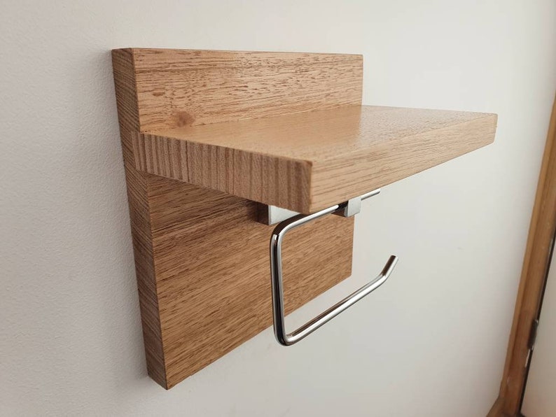 K&ouml;nnte beinhalten: Ein Holzregal mit einem verchromten Toilettenpapierhalter. Das Regal ist aus hellbraunem Holz mit nat&uuml;rlicher Maserung.