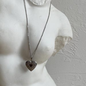Op de afbeelding: Een zilverkleurige hartvormige medaillonketting met een fijne ketting. Het medaillon heeft een ingewikkeld ontwerp en een kleine hartvormige uitsparing. De ketting wordt getoond op een witte buste.