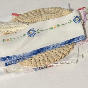 May include: A collection of vintage textiles including a white fabric strip with blue and yellow floral embroidery, a beige crocheted doily, and various white lace trims. The embroidered strip features blue daisy-like flowers and green stems.