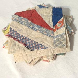 May include: A stack of square fabric quilt blocks featuring a patchwork design with various patterns and colors including red, blue, and white. The blocks are stitched with visible quilting lines and have frayed edges, showcasing a mix of floral and geometric prints.