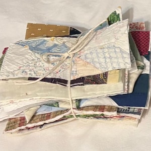 May include: A stack of assorted fabric squares, tied with twine. The squares feature various patterns and colors, including floral, geometric, and paisley designs. The fabrics are a mix of cotton and other textiles, creating a patchwork aesthetic.