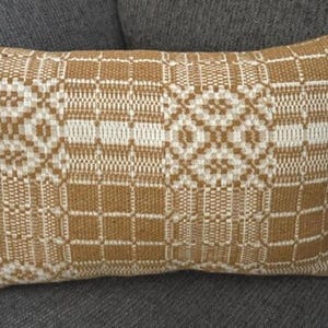 May include: A rectangular throw pillow featuring a woven pattern in shades of mustard yellow and off-white. The design consists of alternating geometric blocks with intricate grid and diamond motifs, creating a textured appearance across the surface.