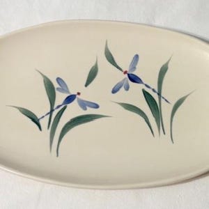 May include: Cream-colored oval serving platter with a hand-painted dragonfly design. The dragonflies are blue with red accents, and the leaves are green. A decorative piece for serving food.