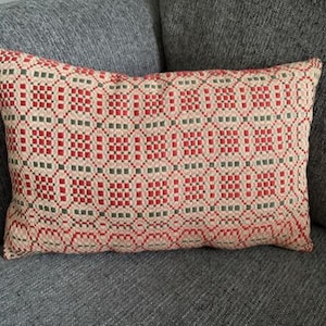 May include: Rectangular decorative pillow with a geometric pattern in red, green, and cream. The pillow is placed on a gray sofa. The pattern consists of squares and circles.