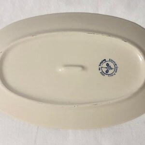 May include: Off-white oval serving platter with a slightly raised rim. The underside features a blue emblem with a dragonfly design and text. A small handle is centered on the underside.