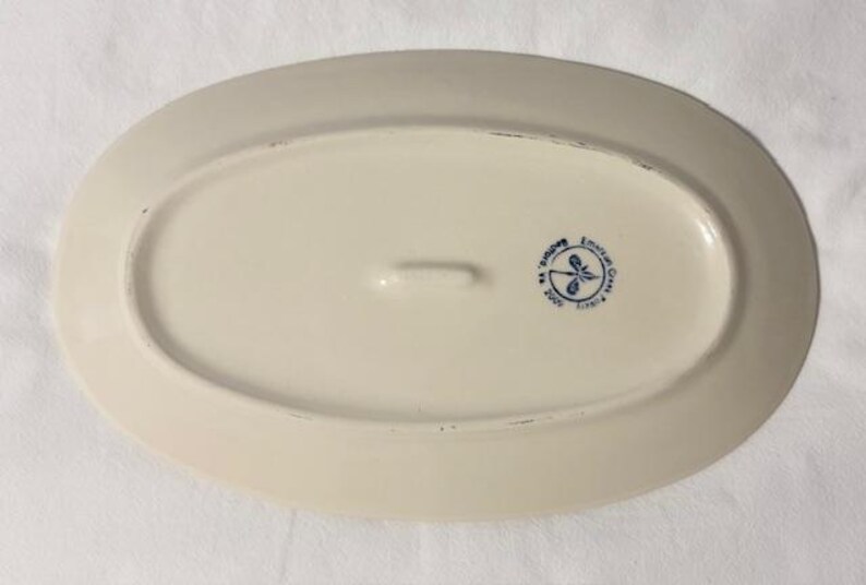 May include: Off-white oval serving platter with a slightly raised rim. The underside features a blue emblem with a dragonfly design and text. A small handle is centered on the underside.