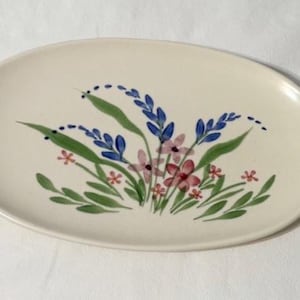May include: Oval ceramic serving platter with a cream-colored base. The platter is decorated with a hand-painted floral design featuring blue, pink, and red flowers with green leaves. The design is centered on the platter.