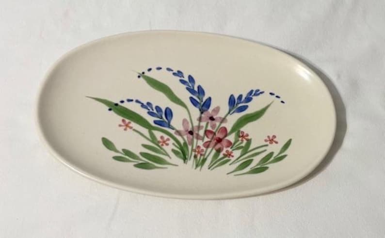 May include: Oval ceramic serving platter with a cream-colored base. The platter is decorated with a hand-painted floral design featuring blue, pink, and red flowers with green leaves. The design is centered on the platter.