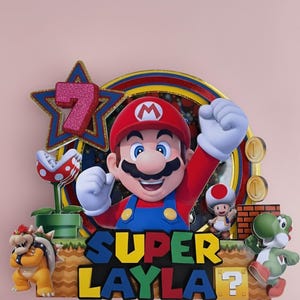 Może przedstawiać: Kolorowa dekoracja na tort w stylu Super Mario, przedstawiająca Mario z uniesioną pięścią, gwiazdą z numerem 7 i imieniem "Layla". Zawiera inne postacie i elementy z gry.