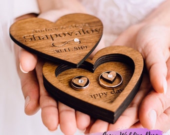 May include: A wooden heart-shaped ring box with the inscription "Audrey & Alex Shapovalov 30.11.2019" and the word "Happy Day" engraved on the inside. Two gold rings with small diamonds are inside the box.