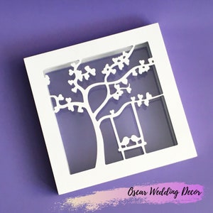May include: White square wall art featuring a tree and swing design. The tree has heart-shaped leaves, and the swing holds two birds. The frame is white, set against a purple background. The text "Oscar Wedding Decor" is at the bottom.
