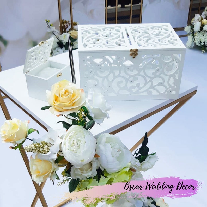 May include: White wedding card box with intricate cut-out designs and a gold latch. The box is on a gold table with white flowers.
