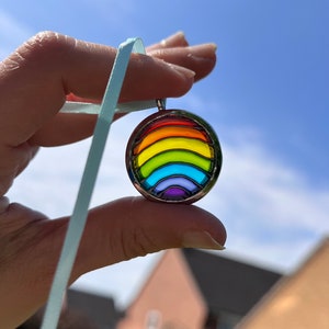 May include: A round pendant with a rainbow design, featuring red, orange, yellow, green, blue, and purple stripes. The pendant is held by a light blue ribbon against a blue sky background. The pendant is a piece of jewelry.
