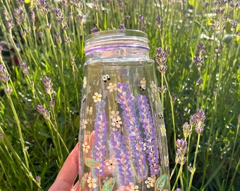 May include: A clear glass jar with a purple, green, and white floral design painted on the side. The jar is filled with lavender flowers and bees.
