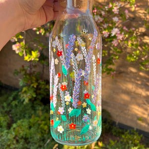 Puede incluir: Una botella de vidrio transparente con un tapón de corcho está pintada a mano con un diseño floral. La botella presenta flores moradas, rojas, amarillas y blancas con hojas verdes.