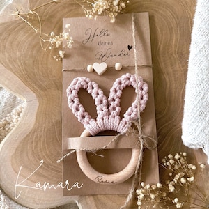 Könnte beinhalten: Ein rosa gehäkeltes Hasenrassel mit einem Holzring. Der Ring ist mit dem Namen "Rosie" graviert. Die Verpackung enthält eine Karte mit dem Text "Hallo kleines Wunder" und zwei kleine Holzh Herzen.