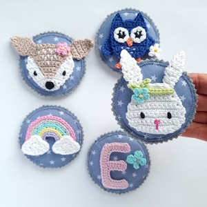 Könnte beinhalten: Fünf gehäkelte Filzpatches mit blauem Sternenhintergrund. Die Patches zeigen einen Regenbogen, ein Reh, eine Eule, ein Kaninchen und den Buchstaben E.