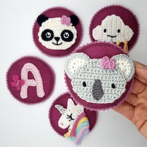 Könnte beinhalten: Fünf Filz-Kreise mit gehäkelten Applikationen. Die Applikationen umfassen einen Panda, eine Wolke, einen Koala, ein Einhorn und den Buchstaben A. Die Kreise sind in einem tiefen Burgunderrot.