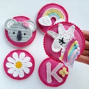 Könnte beinhalten: Fünf rosa Filz-Kreise mit gehäkelten Designs. Die Designs beinhalten einen Koala, eine Gänseblümchen, ein Einhorn, einen Regenbogen und den Buchstaben K mit einem Schmetterling.