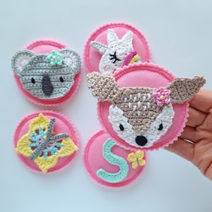 Könnte beinhalten: Fünf rosa Filz-Kreise mit gehäkelten Tier- und Blumenapplikationen. Die Applikationen umfassen einen Koala, ein Einhorn, ein Reh, einen Schmetterling und einen Buchstaben S.