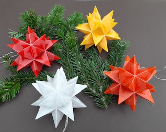 May include: Four paper star ornaments in red, orange, yellow, and white. The stars are arranged on a bed of green pine branches.