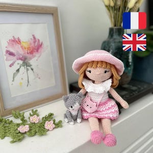 Può includere: Una bambola fatta all'uncinetto con capelli castano chiaro, che indossa un cappello da sole rosa, un colletto bianco con volant e una gonna rosa a motivi con una piccola borsa a tracolla abbinata. Accanto alla bambola c'è un piccolo gatto all'uncinetto grigio e bianco.