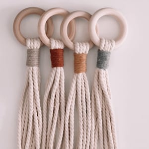 May include: Four macrame plant hangers with wooden rings and different colored accents. The hangers are made of natural cotton rope and have a rustic, bohemian style.