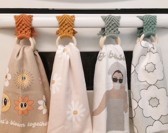 May include: Four kitchen towels hanging on a white oven door handle. The towels are made of white fabric with different patterns. The first towel has a yellow sun and flowers, the second has white daisies, the third has the text "Get Busy" and a woman with sunglasses, and the fourth has large white flowers.