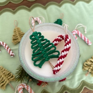 May include: A close-up of handmade Christmas ornaments. Two green yarn Christmas trees and a red and white candy cane ornament are displayed in a white bowl. The ornaments are made with yarn and are tied with twine.