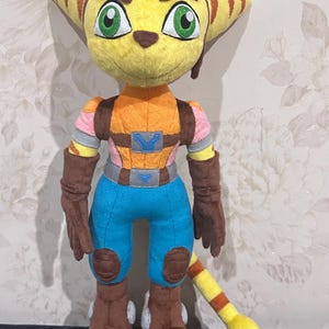 May include: A plush toy of a yellow and orange cat-like character with brown boots, blue pants, and a brown vest. The character has a yellow tail with orange stripes.