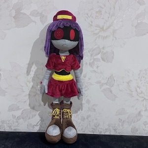 以下が含まれることがあります： 紫色の髪、赤と黄色の服を着たロボットの女の子のぬいぐるみ。おもちゃは暗い表面の上に立っています。
