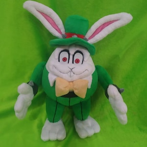 Pode incluir: Um coelho de pelúcia verde com rosto branco e olhos vermelhos usando um chapéu verde com uma faixa vermelha. O coelho tem uma gravata borboleta amarela e está em pé em uma superfície verde.