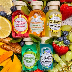 May include: Six small bottles of shampoo with colorful labels and fruit-themed names. The bottles are arranged on a bed of fruit, including strawberries, grapes, lemons, and dragon fruit.