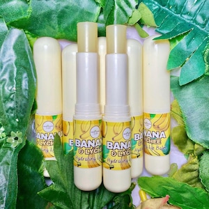 May include: Several lip balm tubes with yellow and white packaging are arranged against a backdrop of green leaves. The labels read "Banana & Lychee Hydrating Lip Balm" with a banana graphic. The lip balm tubes are open, revealing the product.