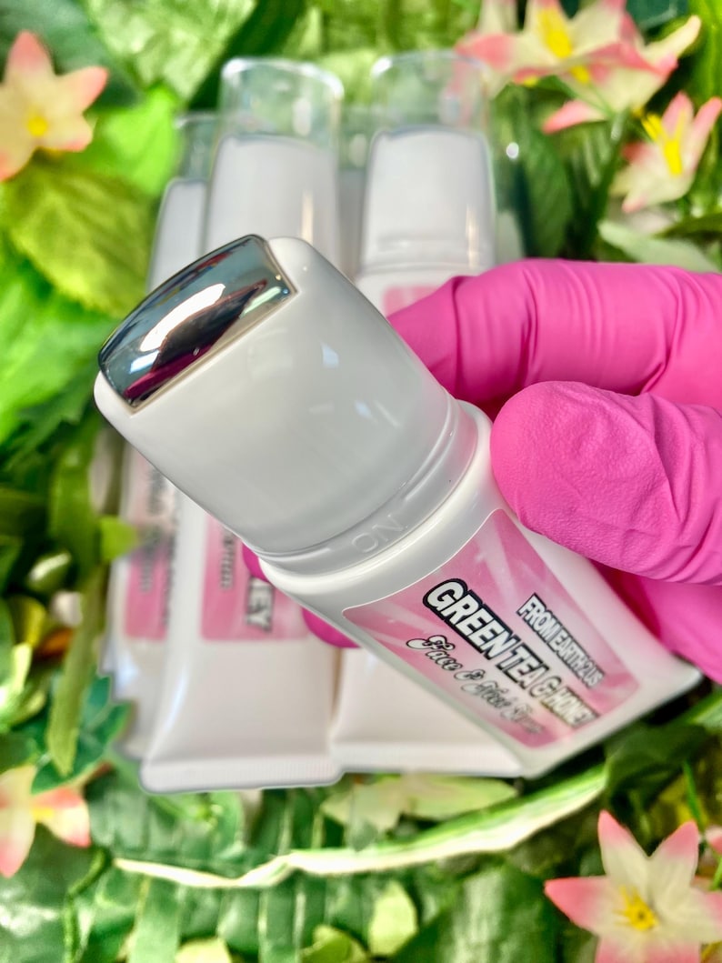 May include: White tubes of Green Tea & Honey face cream with a silver rollerball applicator. The tubes have pink labels with black text. The background includes green foliage and pink flowers.
