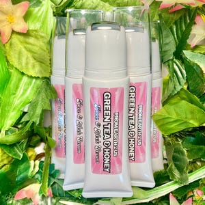 May include: Several white tubes of face and neck serum with pink labels and the text "GREEN TEA & HONEY" and "Face & Neck Serum". The tubes are surrounded by green leaves and pink and yellow flowers.