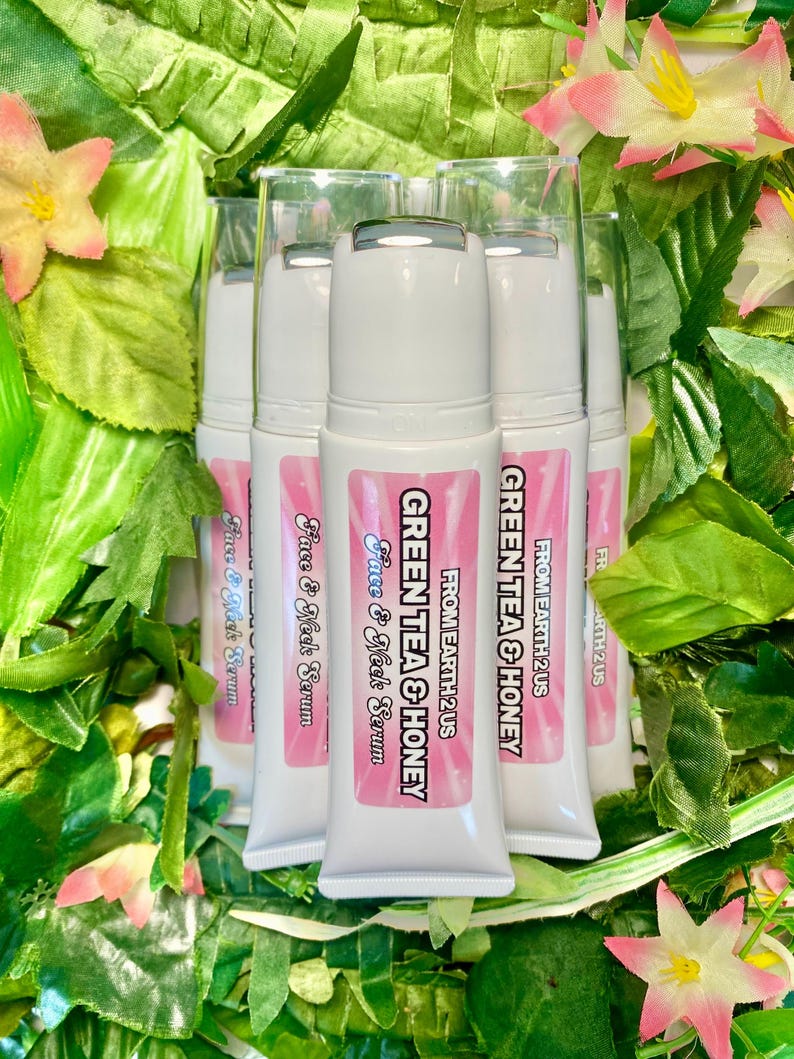 May include: Several white tubes of face and neck serum with pink labels and the text "GREEN TEA & HONEY" and "Face & Neck Serum". The tubes are surrounded by green leaves and pink and yellow flowers.