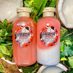 May include: Two bottles of Coconut Hibiscus shampoo and conditioner with a floral label design. The shampoo is a solid pink color, while the conditioner is pink and white. The bottles are surrounded by green leaves and coconut pieces.