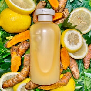May include: A clear glass bottle with a white cap filled with a yellow liquid. The bottle is surrounded by lemon slices and turmeric root pieces on a green leafy background.