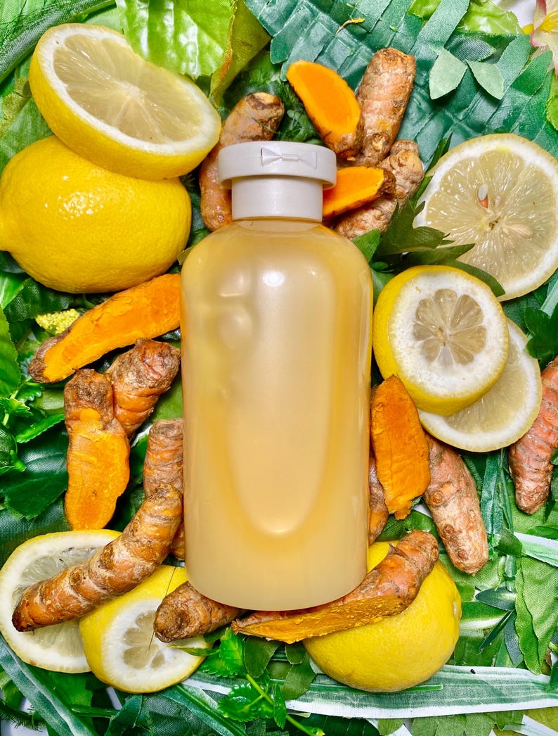 May include: A clear glass bottle with a white cap filled with a yellow liquid. The bottle is surrounded by lemon slices and turmeric root pieces on a green leafy background.