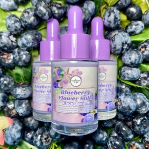 May include: Three glass bottles of Blueberry Flower Milk Face Serum with purple droppers, surrounded by fresh blueberries and green leaves. The bottles have labels with the product name and floral illustrations.