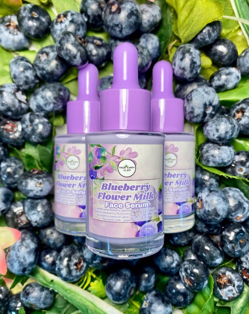 May include: Three glass bottles of Blueberry Flower Milk Face Serum with purple droppers, surrounded by fresh blueberries and green leaves. The bottles have labels with the product name and floral illustrations.
