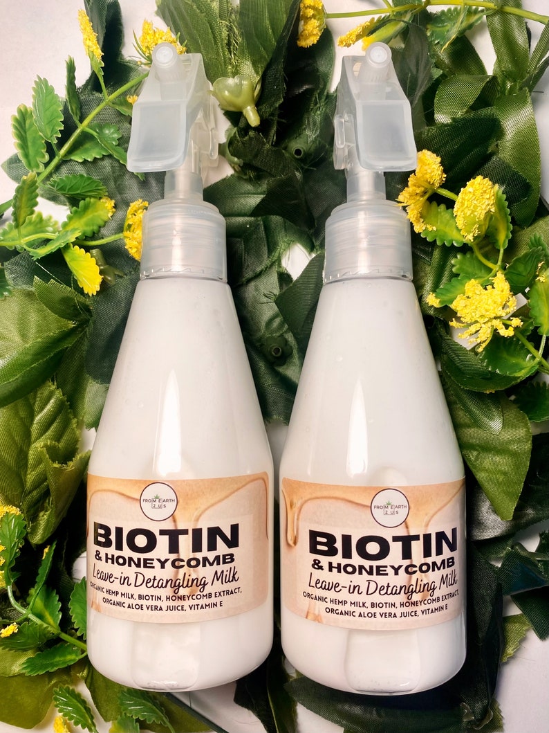 May include: Two white plastic bottles with spray tops, labeled "Biotin & Honeycomb Leave-in Detangling Milk". The bottles are surrounded by green leaves and yellow flowers.