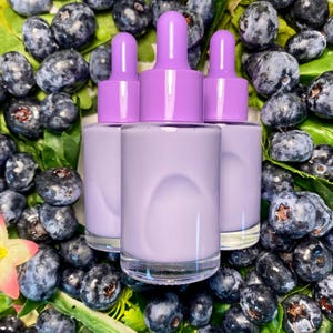 May include: Three lavender-colored dropper bottles with matching caps are surrounded by fresh blueberries and green leaves. The bottles have clear glass bases and are filled with a light purple liquid. The image is a close-up shot, highlighting the product and the natural elements.
