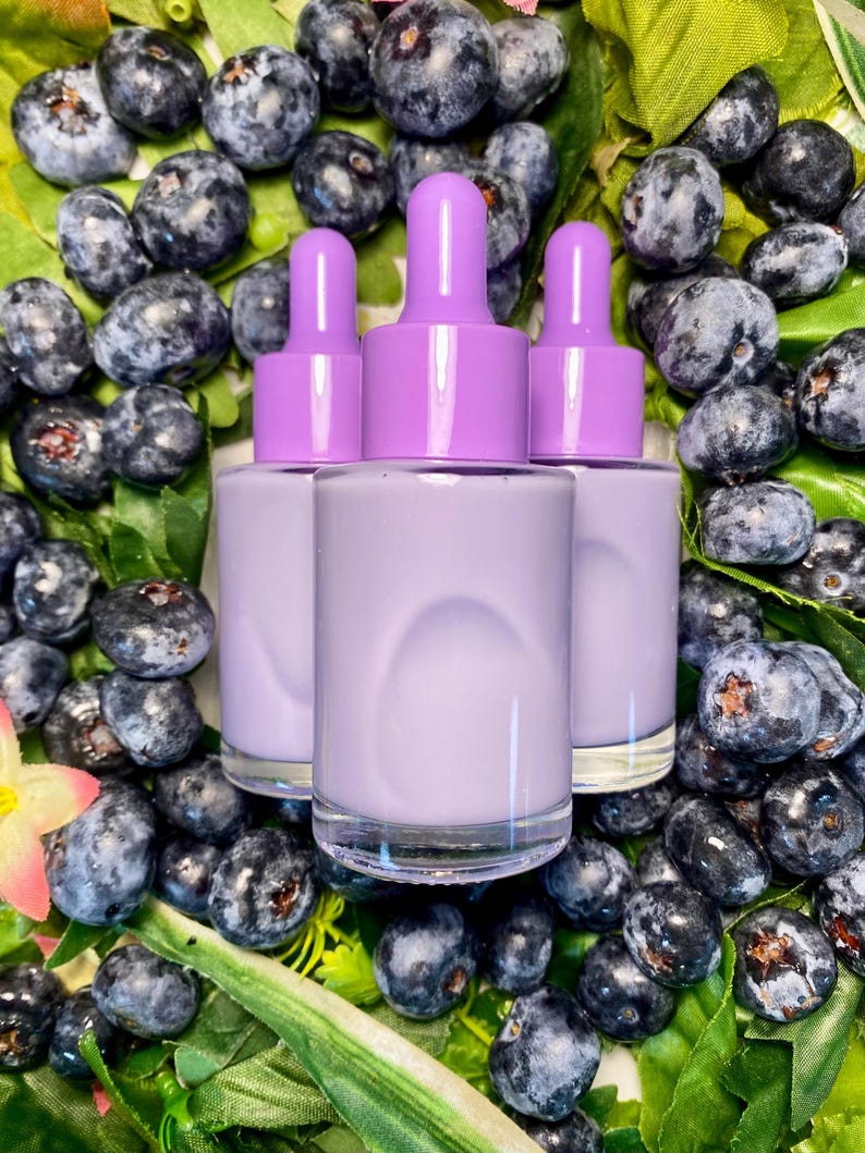 May include: Three lavender-colored dropper bottles with matching caps are surrounded by fresh blueberries and green leaves. The bottles have clear glass bases and are filled with a light purple liquid. The image is a close-up shot, highlighting the product and the natural elements.