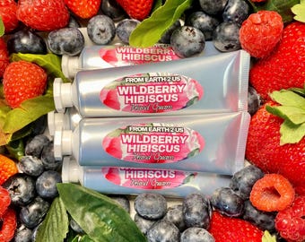 May include: Four tubes of hand cream with the text "Wildberry Hibiscus Hand Cream" are surrounded by fresh strawberries, raspberries, blueberries, and green leaves. The tubes are silver with a pink and white label.