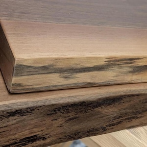 Può includere: Primo piano di due assi di legno sovrapposte. Il legno mostra venature naturali con tonalità marroni chiare e scure. L'asse superiore ha bordi puliti, mentre quella inferiore ha un aspetto più rustico.
