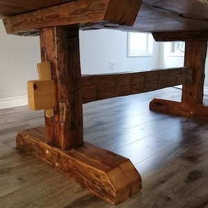 Puede incluir: Una mesa de madera rústica con una tapa gruesa de madera natural y una base de caballete. La base está hecha de vigas gruesas y toscamente labradas que se unen con clavijas de madera. La mesa tiene un acabado natural y desgastado.