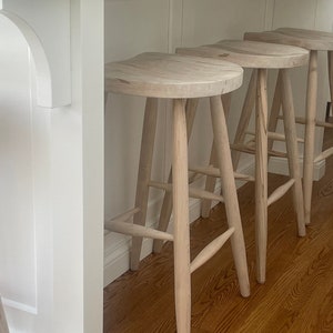 Puede incluir: Tres taburetes de bar de madera blanca con un diseño sencillo. Los taburetes tienen un asiento redondeado y cuatro patas.